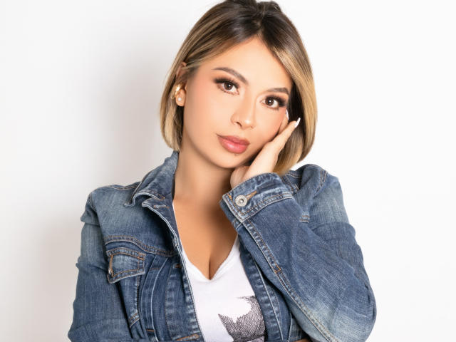 IsaKors with short blonde hair, wearing a denim jacket and white top, resting her cheek on her hand, brown eyes, subtle makeup, pink lips, looking at the camera.