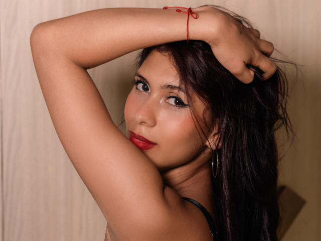 DanikaSerov with dark hair, red lipstick, and hoop earrings, arms raised, showing tan skin and subtle makeup, wearing a red bracelet on her wrist.