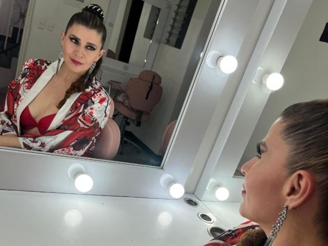 MegganSylph with dark hair in a bun, wearing a red bra and floral robe, looking in a mirror with lights, showing her tits and a confident smile.