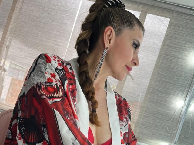 MegganSylph with braided brown hair, red and black floral kimono, large silver earrings, pink lipstick, and a slight smile, looking to the right. Boobs visible through kimono.