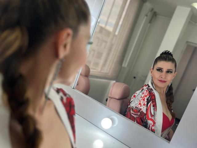 MegganSylph with her back to the mirror, brown hair in a braid, wearing a red and white floral top, looking at her reflection, showing her tits in the mirror.