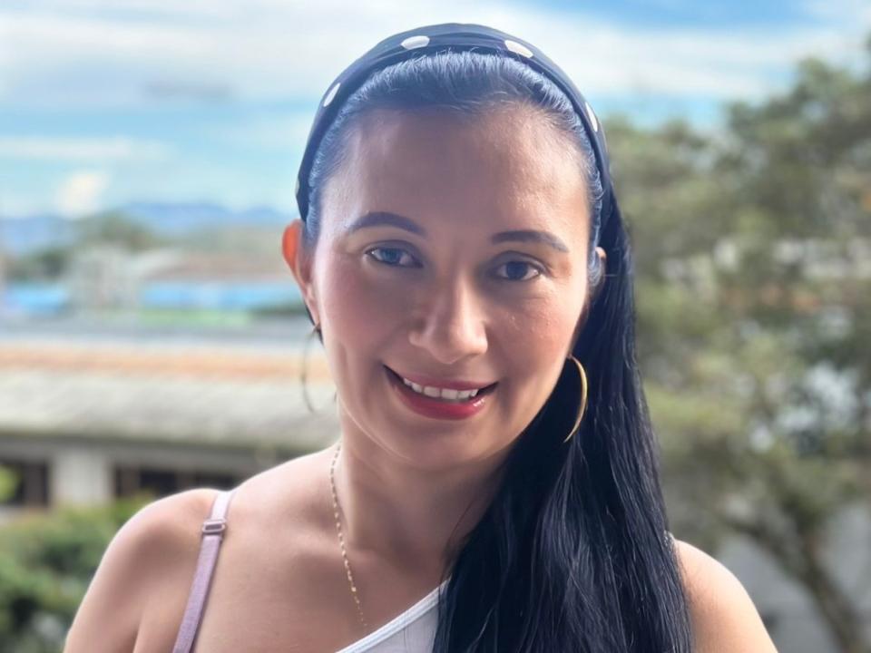 GraceK has dark black hair in a headband, smiling with red lips, wearing a white strap, gold hoop earrings, and a thin necklace, showing her cute face.