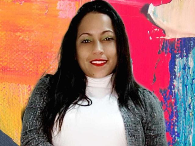 ScarlettBarness has dark hair, red lipstick, and a white turtleneck with a gray cardigan, smiling at the camera. Her ass is not visible.