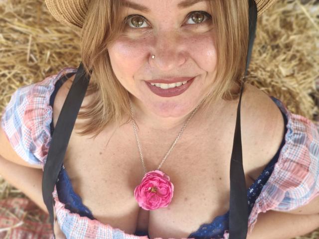PeachyTease-hot with blonde hair, wearing a hat, plaid shirt, and black straps, has pink flower necklace, exposed blue lace bra, showing her big boobs, smiling, nose ring, and dark lipstick.