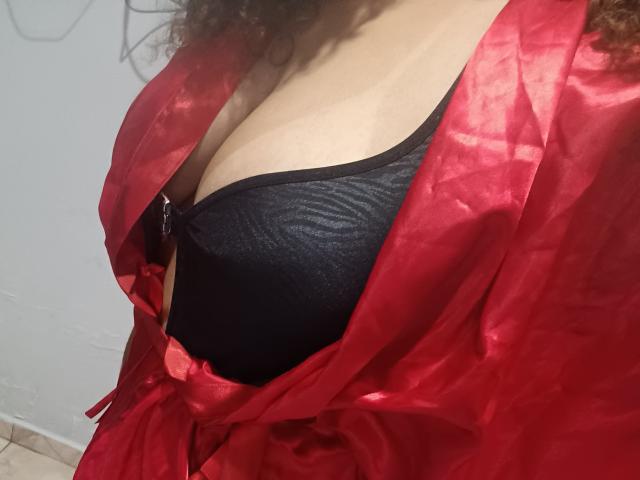 CabelereiraSexy69's red silky robe is open, showing her black bra with her large, rounded tits partially visible. Her curly brown hair is slightly visible.