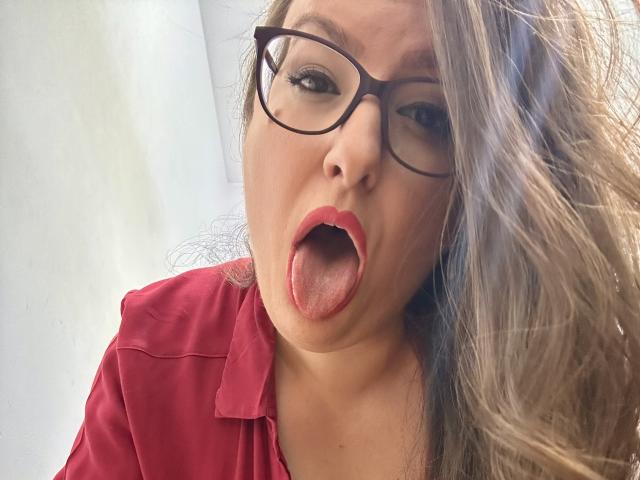 BadAngel with tongue out, red lips, glasses, and long brown hair, wearing a red shirt, has a slightly open mouth and a playful expression.