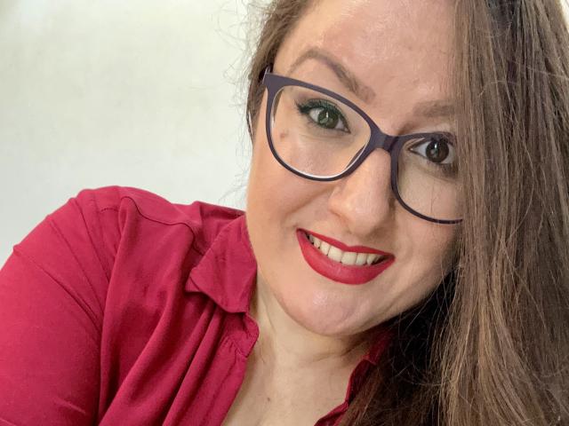 BadAngel has red lipstick, glasses, and a pink shirt, smiling with green eyes, brown hair, and a cute face. Her tits are small, and she's looking at the camera.