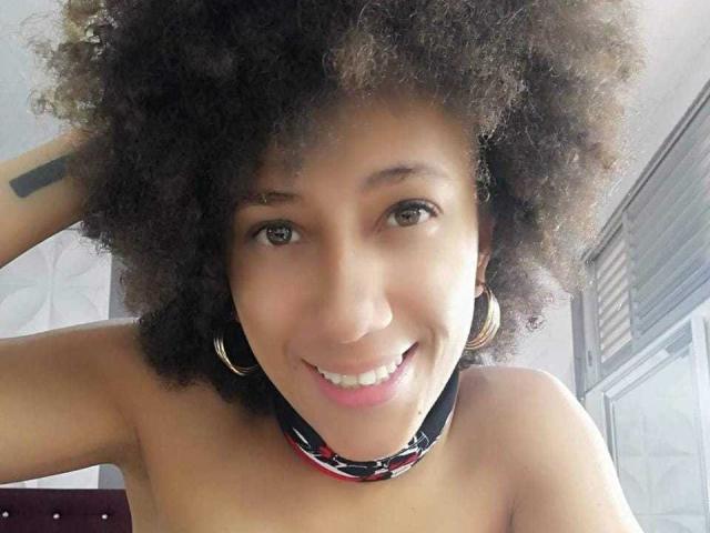 SucyAndrea with curly brown hair, smiling, wearing large hoop earrings and a black necklace with red and white patterns, showing a tattoo on her left arm.