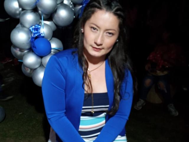 CristallDiaz has long brown hair, blue cardigan, striped top, and a gold necklace, with a cute smile, standing in front of silver balloons and a blue ornament.