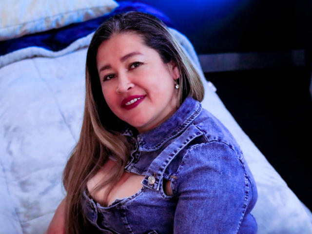 NattaReed with long blonde hair, blue denim shirt, and red lipstick, smiling at the camera, showing her tits in a cleavage cutout, sitting on a white bed.