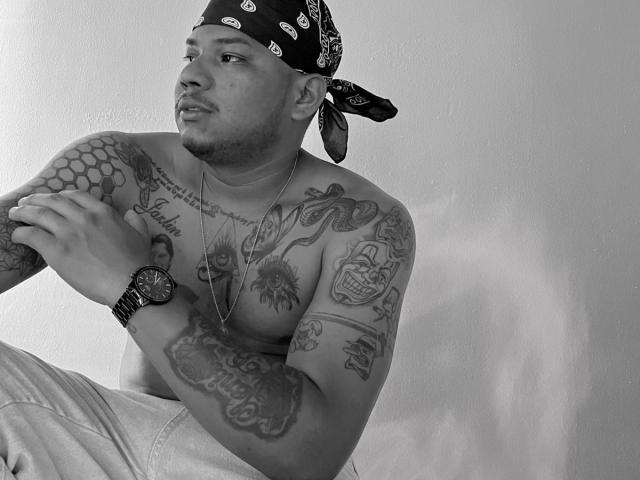SamSexy69, un uomo tatuato con una bandana, senza camicia, che indossa jeans e un orologio, guarda di lato con un'espressione leggermente curiosa. I suoi tatuaggi sulle braccia sono ben visibili..