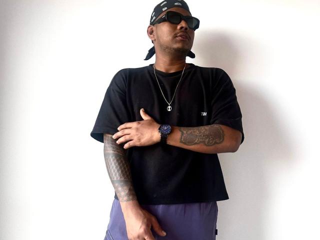 SamSexy69 trägt ein schwarzes T-Shirt, eine violette Hose, eine schwarze Kappe, eine Sonnenbrille, eine Halskette und eine Uhr. Er hat Tattoos, einen Arm, der über den Körper gekreuzt ist, und einen ernsten, leicht schmollenden Gesichtsausdruck..