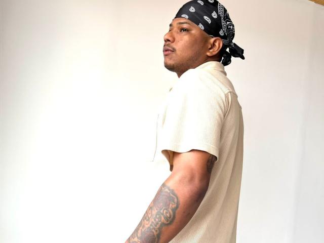 SamSexy69, ein dunkelhäutiger Mann mit einer schwarzen Bandana, einem weißen T-Shirt und einem tätowierten Arm, der zur Seite schaut und ein leicht schmollendes Gesicht hat..