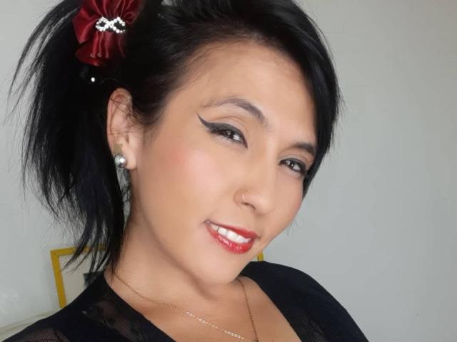 DaniaHot69 hat schwarzes Haar zu einem Pferdeschwanz gebunden, mit einer roten Blume, Eyeliner, Perlenohrringen, rotem Lippenstift und einem schwarzen Spitzenoberteil, und lächelt in die Kamera..