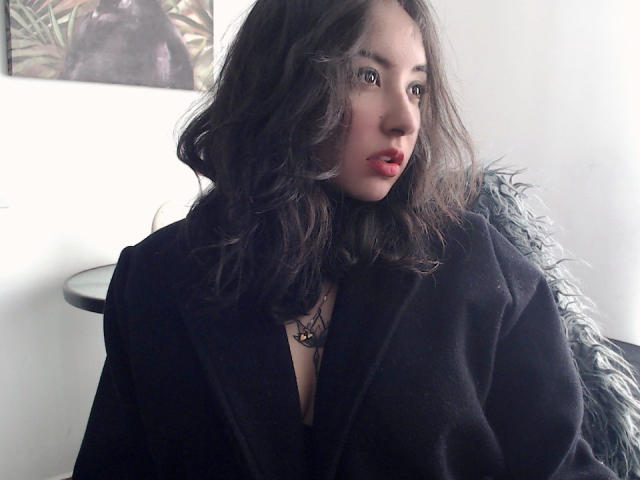 SusanMonroy with wavy dark brown hair, wearing a black robe, red lips, and a necklace with a small pendant, looking to the side, sitting on a fluffy blue chair.