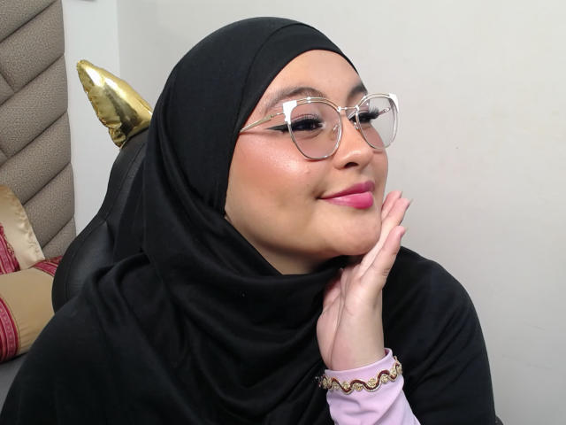HelueJamil, avec son hijab noir, son rouge à lèvres rose, ses lunettes transparentes et son bracelet rose, souriante, le menton dans la main, montrant ses jolies fesses et sa peau éclatante..
