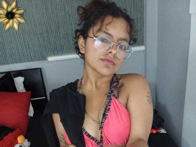 DarcyTear with curly black hair, glasses, pink halter top, black jacket, nose piercing, and tattoos, pouting slightly, in a gray-walled room with red pillows.