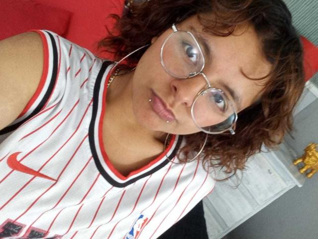 DarcyTear with curly brown hair, wearing glasses, white Nike soccer shirt with red stripes, and a nose piercing, looking at the camera with a slight pout.
