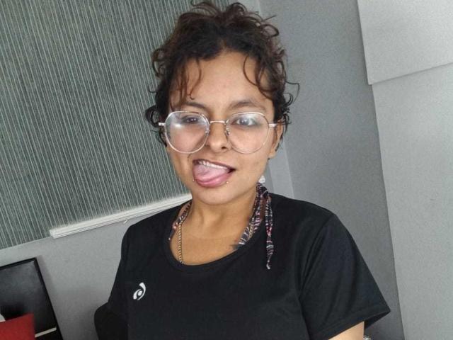 DarcyTear with curly black hair, glasses, and tongue out, wearing a black t-shirt and colorful necklace, has medium brown skin and a playful expression.