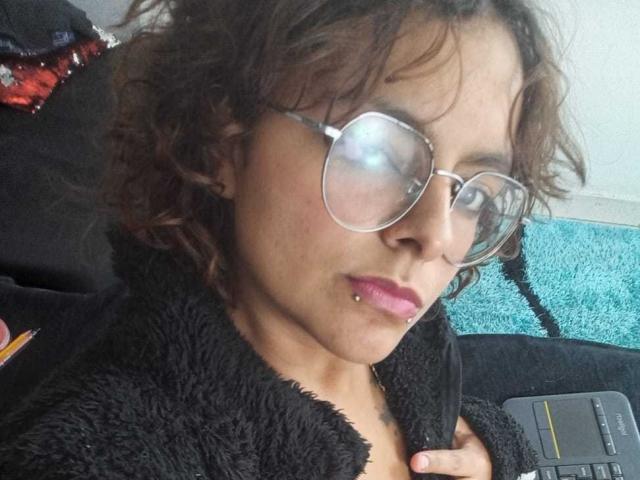 DarcyTear with curly brown hair, wearing glasses, black fuzzy robe, and purple lip ring, showing a pierced lower lip, looking slightly to the side.