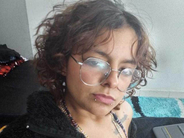 DarcyTear with curly brown hair, glasses, and a nose piercing, wearing a black hoodie, showing a lip piercing, and a tattoo on her shoulder.