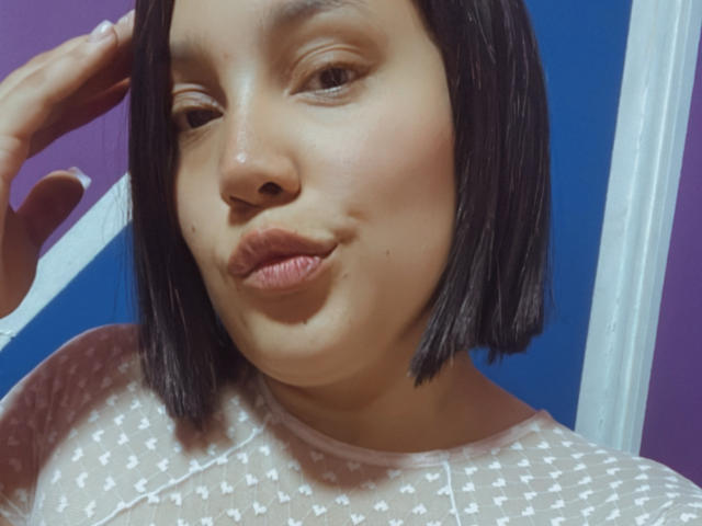 SamantaMuller with a cute pout, short black bob, light brown skin, wearing a sheer white patterned top, touching her head with a hand. Her lips are slightly puckered.