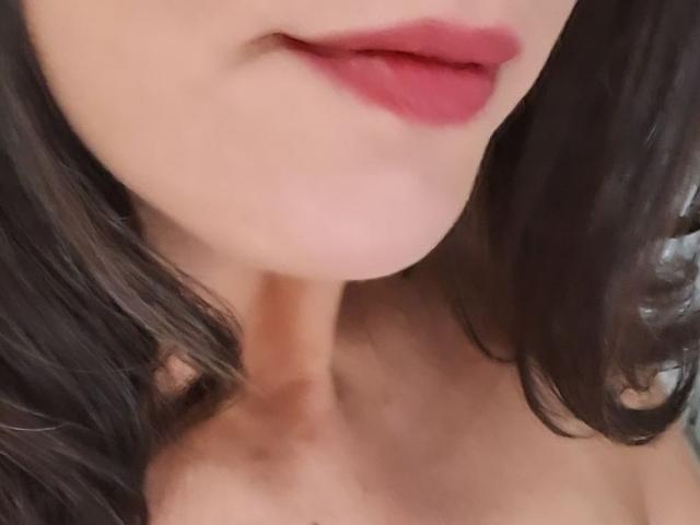 MeahBrunette's face with red lipstick, slightly parted pinkish-red lips, and wavy dark brown hair, showing a soft, fair skin neck. Her ass is not visible.