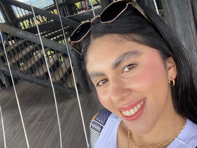 LilyGreay has dark hair, brown eyes, and a pink blush on her face, smiling with red lipstick, wearing sunglasses on her head, and gold earrings with a chain.
