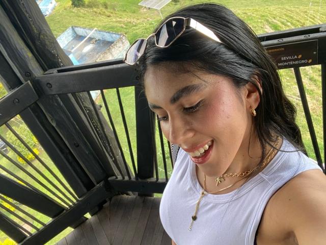 LilyGreay with black hair, white tank top, smiling down, small gold necklace, earrings, sunglasses on head, sitting on black metal balcony, green grass and blue tarp in background.