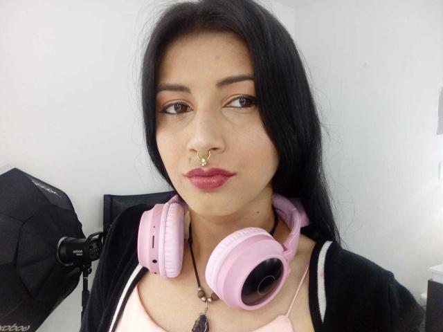 AlisKooper, with long black hair, pink headphones, nose ring, and pink lipstick, wearing a black top, looking at the camera, in a selfie.