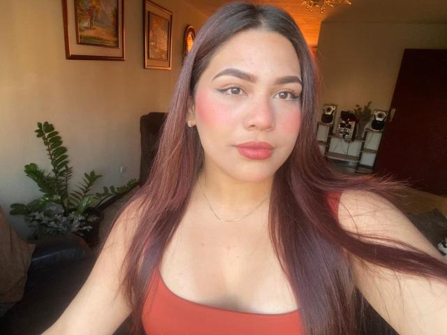 SamanthaLust met lang bruin haar, een rood tanktop en roze blush, die haar borsten en volle, rode lippen laat zien, zittend in een zonnige kamer..