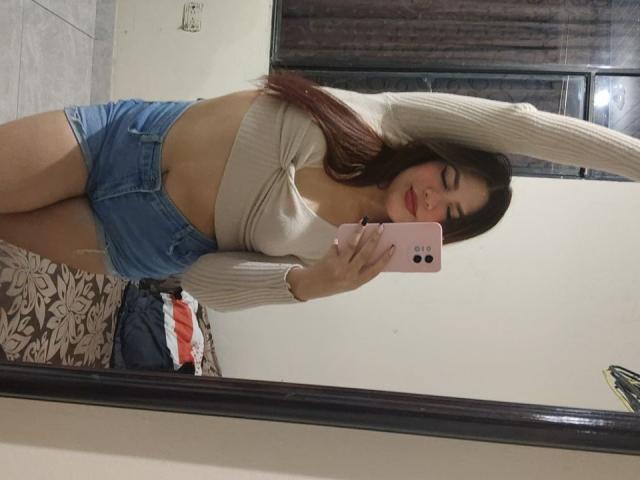 SamanthaLust, avec ses longs cheveux bruns, son rouge à lèvres rouge, son crop top beige et son short en jean bleu, prend un selfie devant un miroir, montrant ses seins et ses fesses, tout en tenant un téléphone rose..