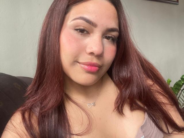 SamanthaLust hat rotbraunes Haar, rosafarbene Lippen und ein leichtes Lächeln. Sie trägt ein graues Oberteil mit einer Herz-Kette und zeigt ihre mittelgroßen Brüste. Ihre Haut ist hell..
