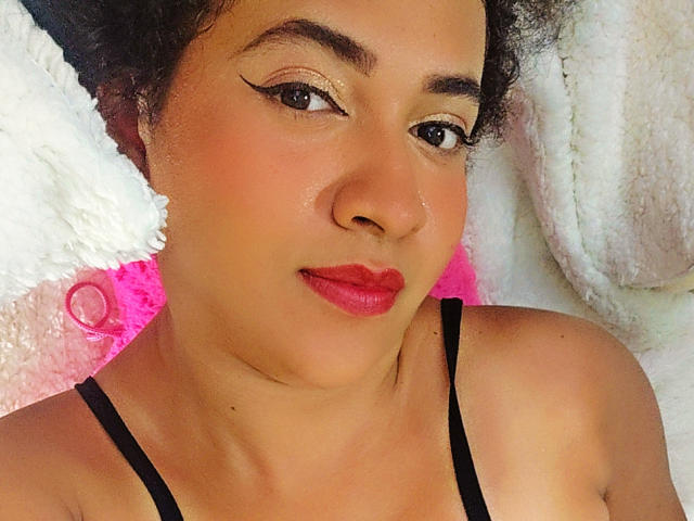 AleWalker has medium brown skin, curly black hair, and red lipstick. She's wearing black straps, has winged eyeliner, and a small nose ring. Her tits are slightly visible.
