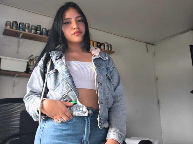 Xianalu mit langen, schwarzen Haaren, die eine Jeansjacke, ein weißes Top und blaue Jeans trägt, steht in einem Raum und hält einen grünen Feuerzeug in der Nähe ihrer Jeans..