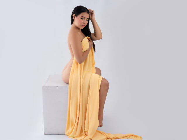 CristalDolce, naked, with long black hair, sits on a white cube, covering her tits and ass with a yellow sheet, looking down, in a minimalist studio.