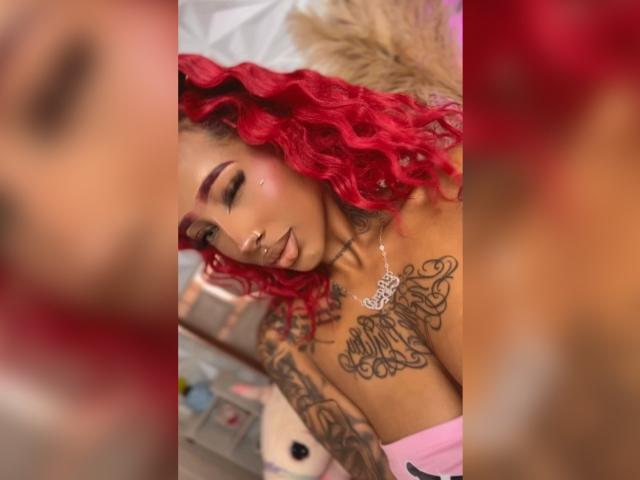 EvanYelin69 with red curly hair, tattoos on chest and arm, nose ring, sleeping with eyes closed, pink top, blurred red and brown edges in the foreground.