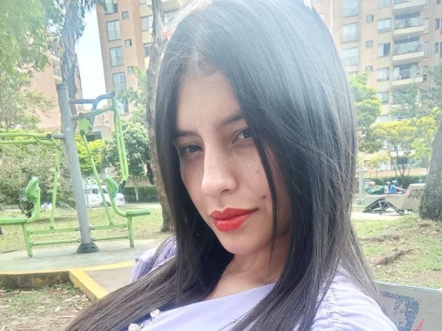 MadissonFontaine with long black hair, red lipstick, and a small mole on her right cheek, wearing a purple shirt, in a park with green benches and a yellow edge.