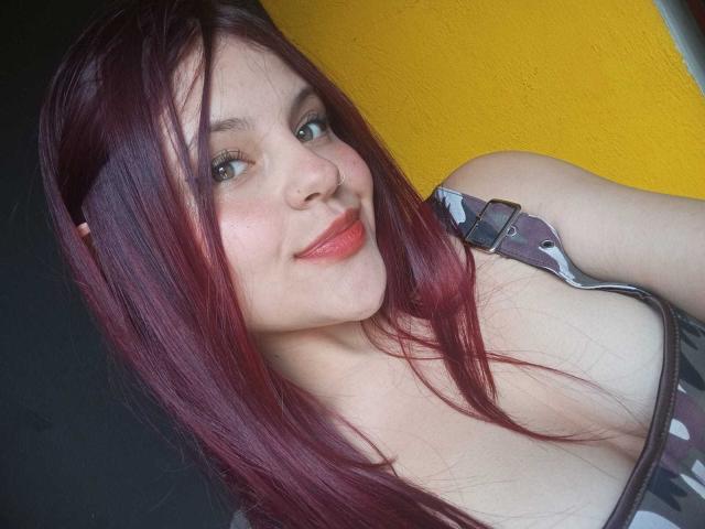 Sophie Hill hat rotes Haar, grüne Augen und lächelt in die Kamera, wobei sie roten Lippenstift trägt. Sie zeigt ihre Brüste in einem Camouflage-Top mit einem Träger über der Schulter..