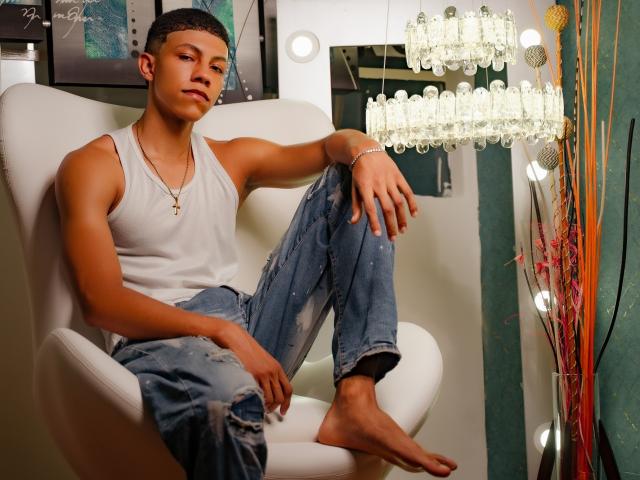 BillySketh, a slim young dude with tan skin and short dark hair, wears a white tank top and ripped jeans, sitting barefoot in a white chair, looking at camera.
