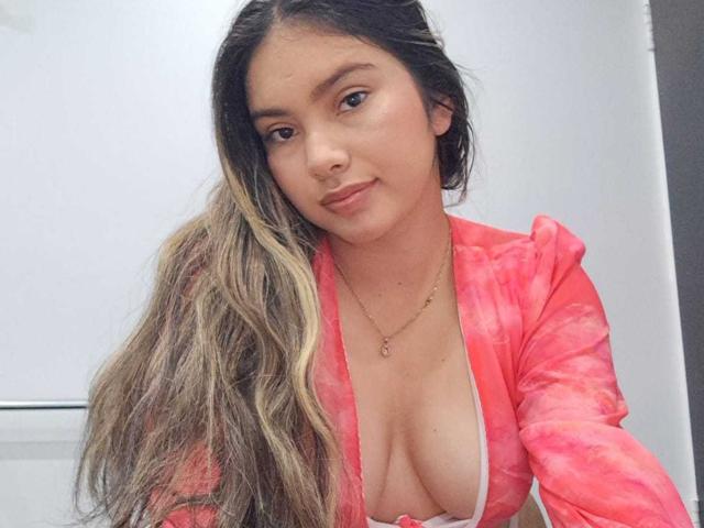 ValeryThomss with long, wavy blonde and brown hair, wearing a pink tie-dye top, exposing her big, perky boobs, and a small, delicate necklace, smiling slightly.