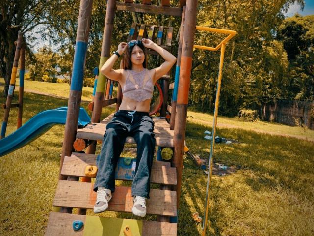 SanzJulie with hands in black hair, wearing a purple lace crop top, black pants, and white sneakers, sitting on a wooden playground structure, blue slide to the left.