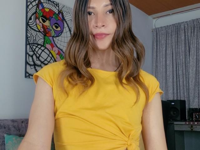 SanzJulie has wavy brown hair, a yellow shirt, and a cute smile. She's showing her tits slightly in the photo. She's standing in a room with gray curtains.