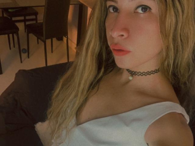 SanzJulie with long blonde hair, wearing a white top, choker, and red lipstick, showing her tits and a small pendant. She's looking at the camera with a serious expression.