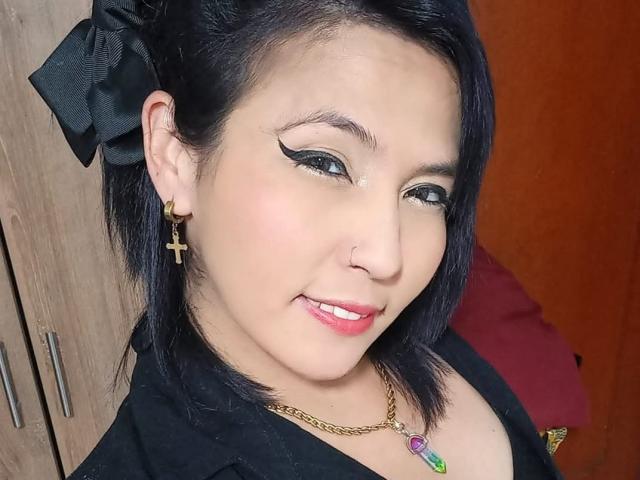 DaniaHot69 has black hair, a black bow, gold cross earrings, winged eyeliner, pink lipstick, a nose ring, and a gold necklace with a purple gem. She's smiling.