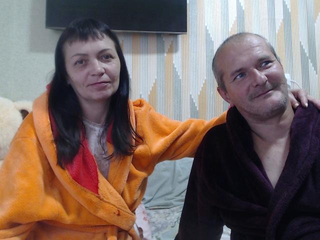 PxoenixAurora, con i capelli scuri, un accappatoio arancione e una fodera rossa, seduta accanto a PxoenixAurora calva, con un accappatoio viola scuro, entrambe sorridenti, in una camera da letto..