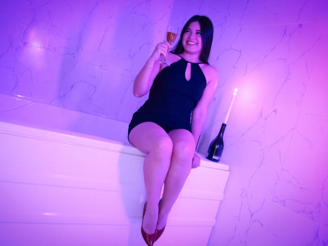 CamilaSofiia in a black halter dress, sitting on a white bathtub, holding a glass of red wine, smiling, with red heels, and a bottle on the right.