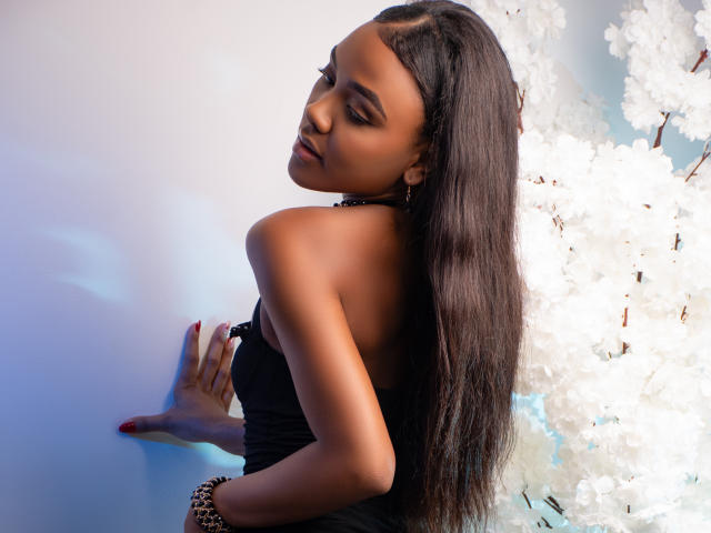AryJonnes with long black hair, dark skin, and a black strapless top, looking back with eyes closed, touching wall with one hand, white cherry blossoms in background.