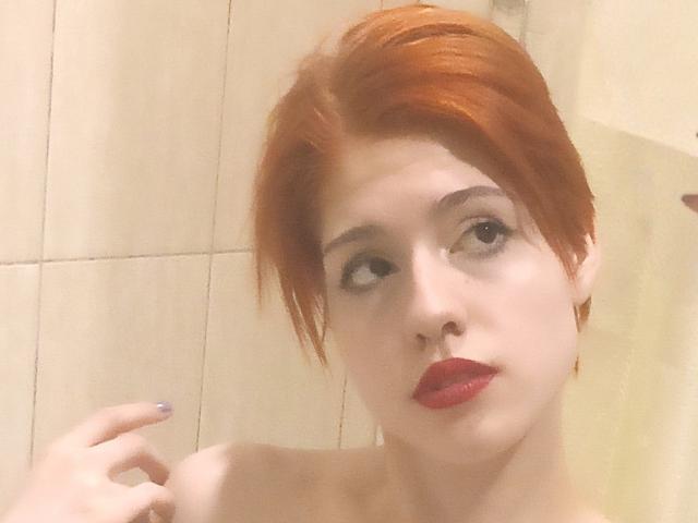 LadyIlona with red hair, pale skin, and red lipstick, looking to the side with a slight smirk, showing her bare shoulders and purple nail polish on her right hand.