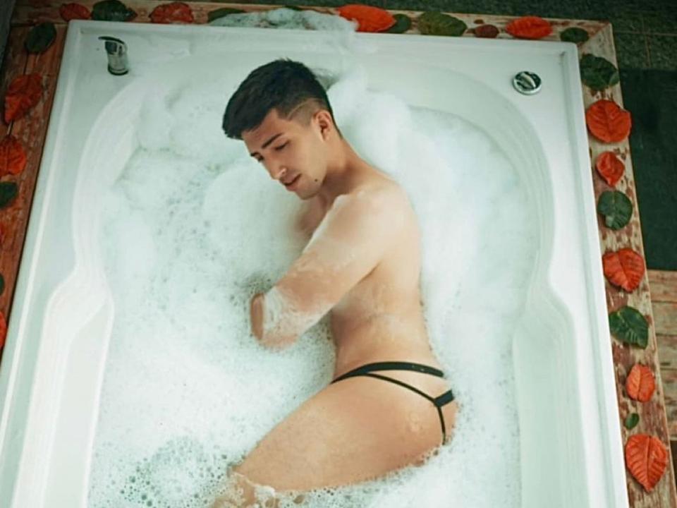 Een aantrekkelijke vrouw, met een strakke bil, draagt een zwarte string en zit in een bruisend wit bad, met haar armen over elkaar, naar beneden kijkend, omringd door schuimende bubbels..